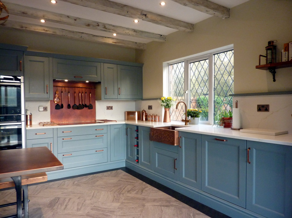 Large traditional galley kitchen/diner in West Midlands with a belfast sink, beaded cabinets, blue cabinets, quartz worktops, orange splashback, black appliances, vinyl flooring, an island, grey floors, white worktops and exposed beams.