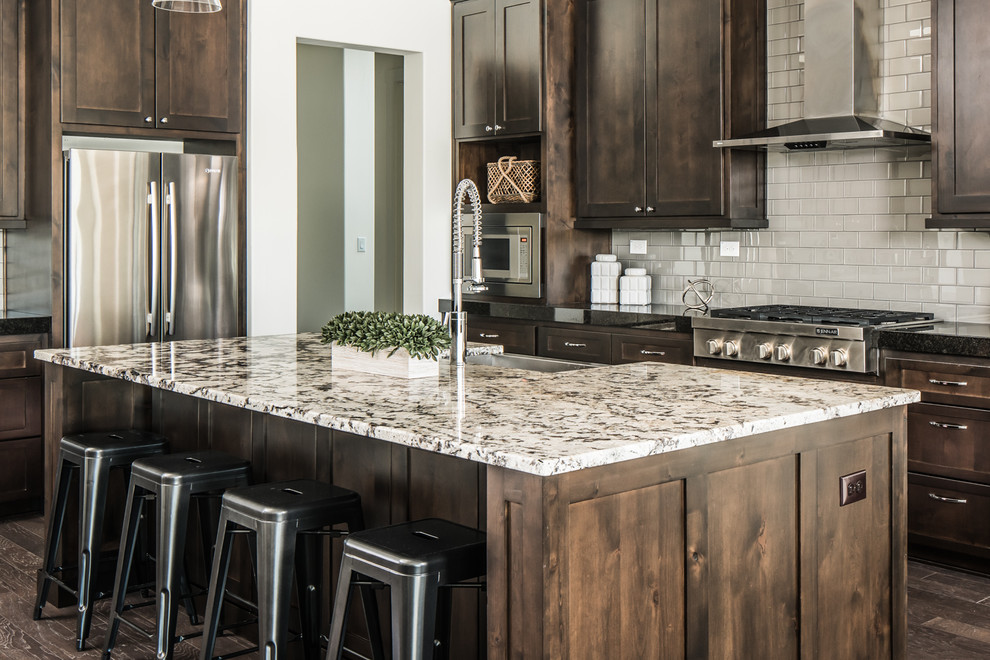 This is an example of a large transitional l-shaped eat-in kitchen in Austin with flat-panel cabinets, brown cabinets, granite benchtops, grey splashback, glass tile splashback, stainless steel appliances, with island, a farmhouse sink and dark hardwood floors.