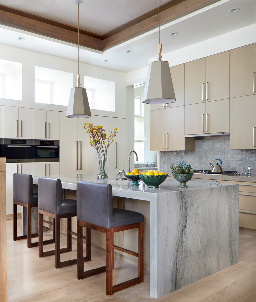 Design ideas for a large contemporary kitchen in Denver with flat-panel cabinets, stone tile splashback, stainless steel appliances, light hardwood floors, with island, beige cabinets and grey splashback.