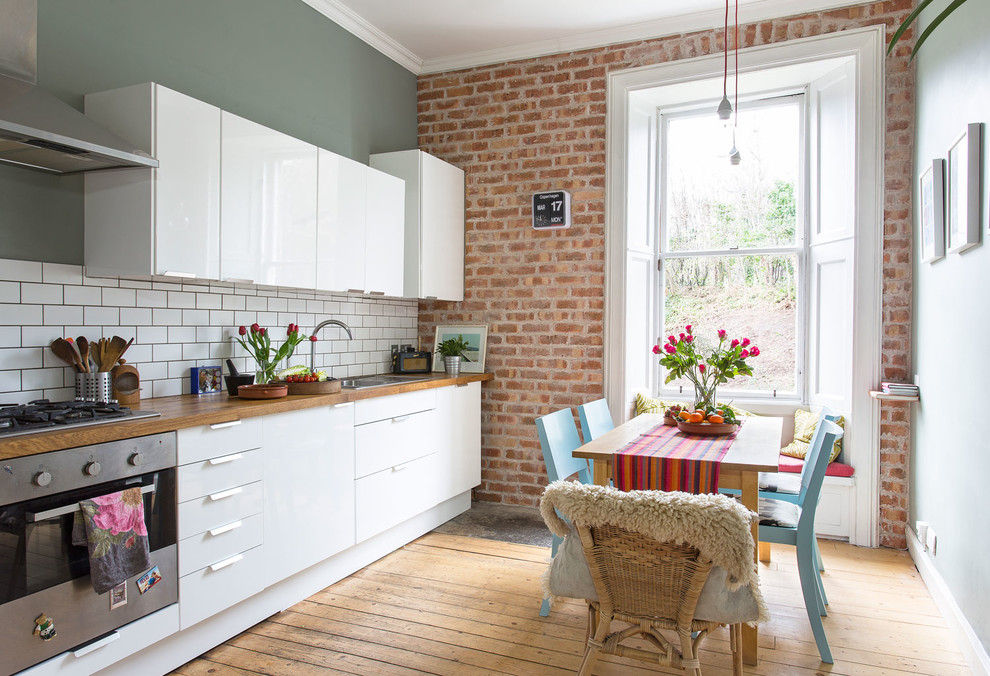 Photo of an eclectic single-wall eat-in kitchen in Edinburgh with a drop-in sink, flat-panel cabinets, white cabinets, white splashback, subway tile splashback, stainless steel appliances, medium hardwood floors, no island, brown floor, wood benchtops and brown benchtop.