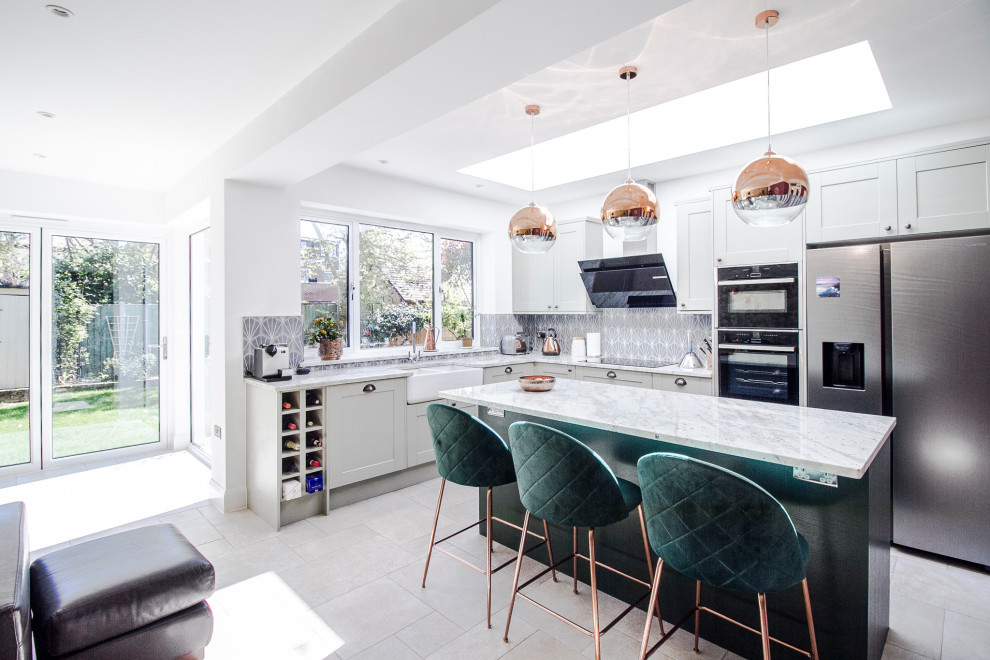 Mid-sized transitional l-shaped open plan kitchen in London with a farmhouse sink, shaker cabinets, granite benchtops, blue splashback, ceramic splashback, stainless steel appliances, limestone floors, with island, grey floor and white benchtop.