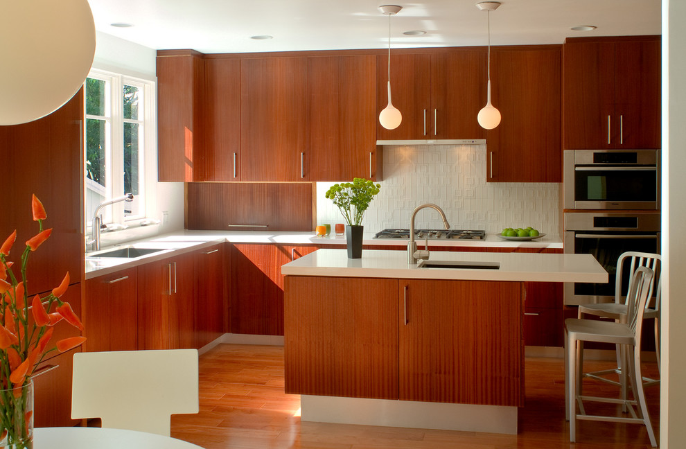 This is an example of a midcentury l-shaped kitchen in Los Angeles with flat-panel cabinets, medium wood cabinets, with island, a single-bowl sink, solid surface benchtops, white splashback, ceramic splashback, stainless steel appliances and medium hardwood floors.