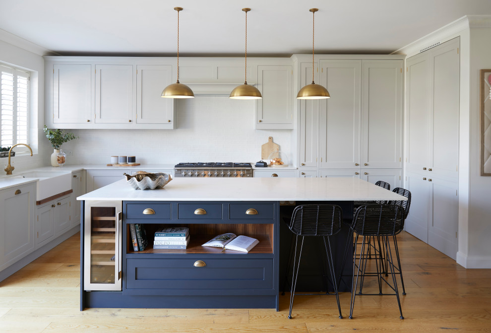 Photo of a transitional l-shaped kitchen in London with a farmhouse sink, shaker cabinets, grey cabinets, white splashback, stainless steel appliances, medium hardwood floors, with island, brown floor and white benchtop.