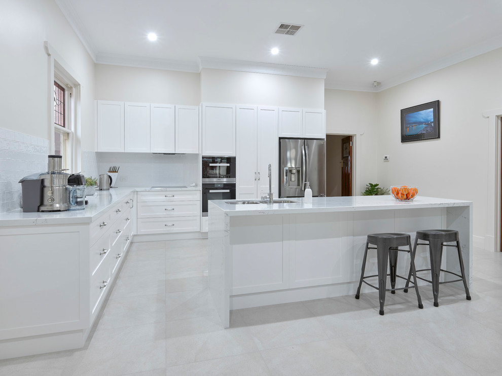 This is an example of a large contemporary l-shaped open plan kitchen in Other with a double-bowl sink, shaker cabinets, white cabinets, quartz benchtops, white splashback, ceramic splashback, stainless steel appliances, porcelain floors, with island and white benchtop.