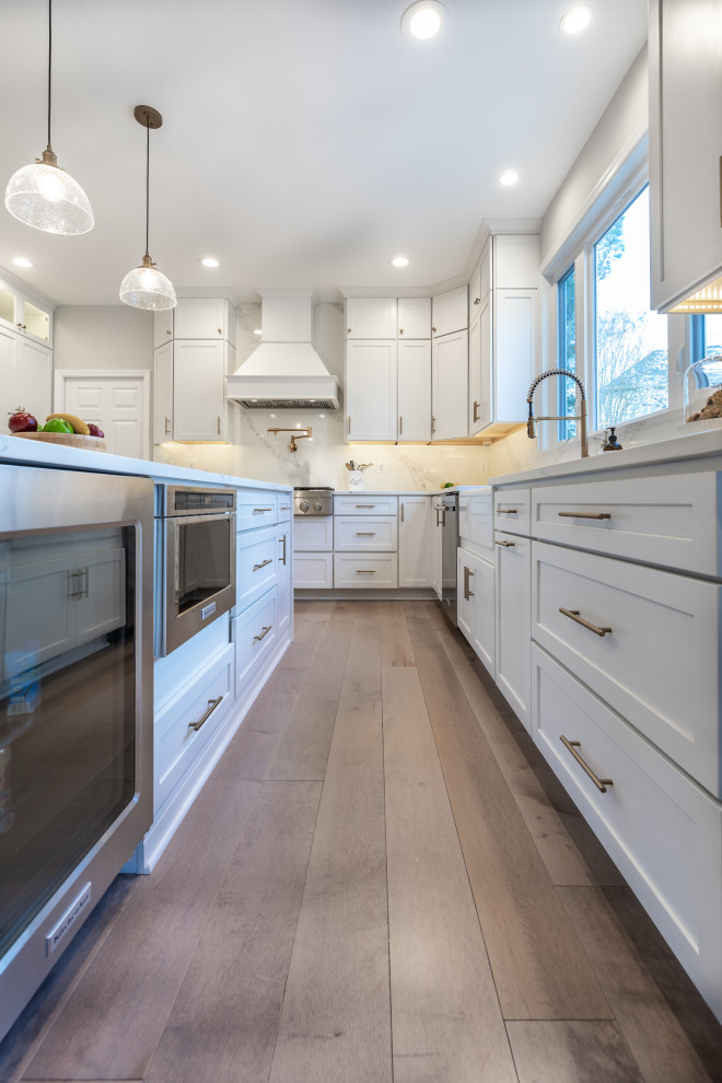 Design ideas for a medium sized contemporary l-shaped kitchen/diner in DC Metro with a belfast sink, beaded cabinets, white cabinets, quartz worktops, white splashback, stainless steel appliances, light hardwood flooring, an island, brown floors and white worktops.