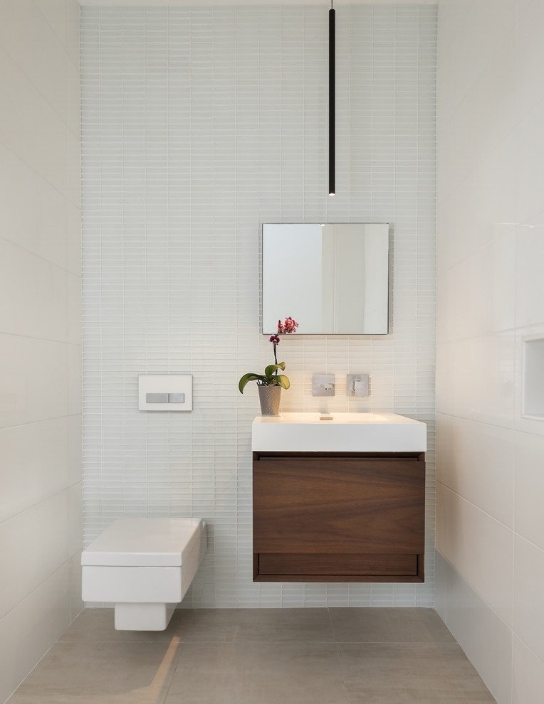 Inspiration for a mid-sized powder room in Phoenix with flat-panel cabinets, dark wood cabinets, a wall-mount toilet, gray tile, white tile, porcelain tile, white walls, porcelain floors, an integrated sink, solid surface benchtops, beige floor and white benchtops.