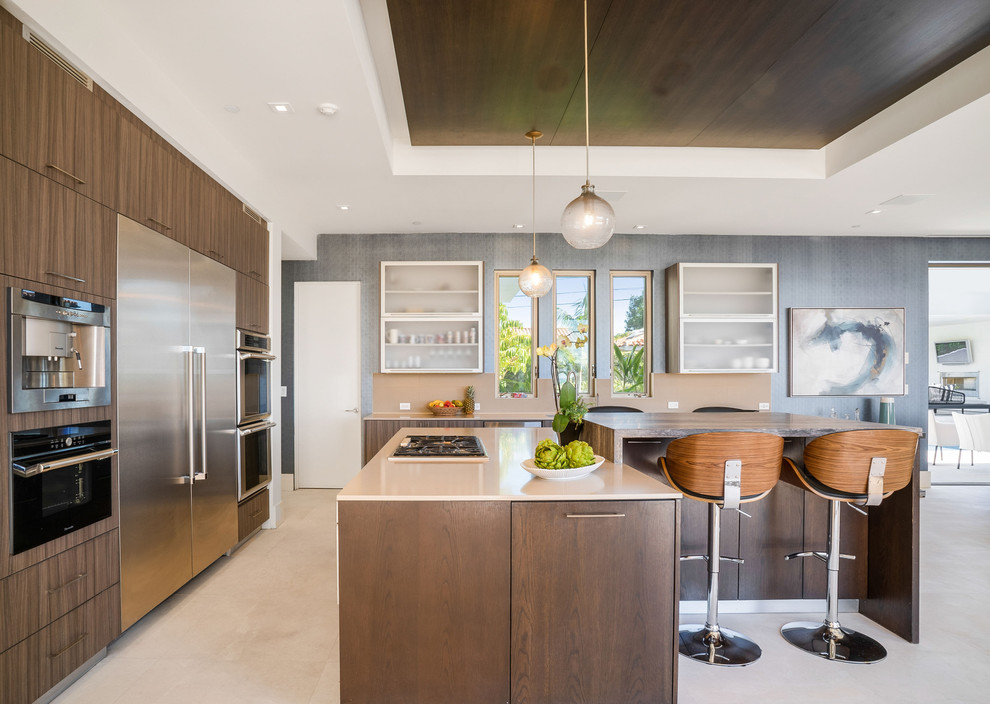 Large beach style open plan kitchen in Los Angeles with flat-panel cabinets, solid surface benchtops, beige splashback, stainless steel appliances, travertine floors, multiple islands, beige benchtop, dark wood cabinets and beige floor.
