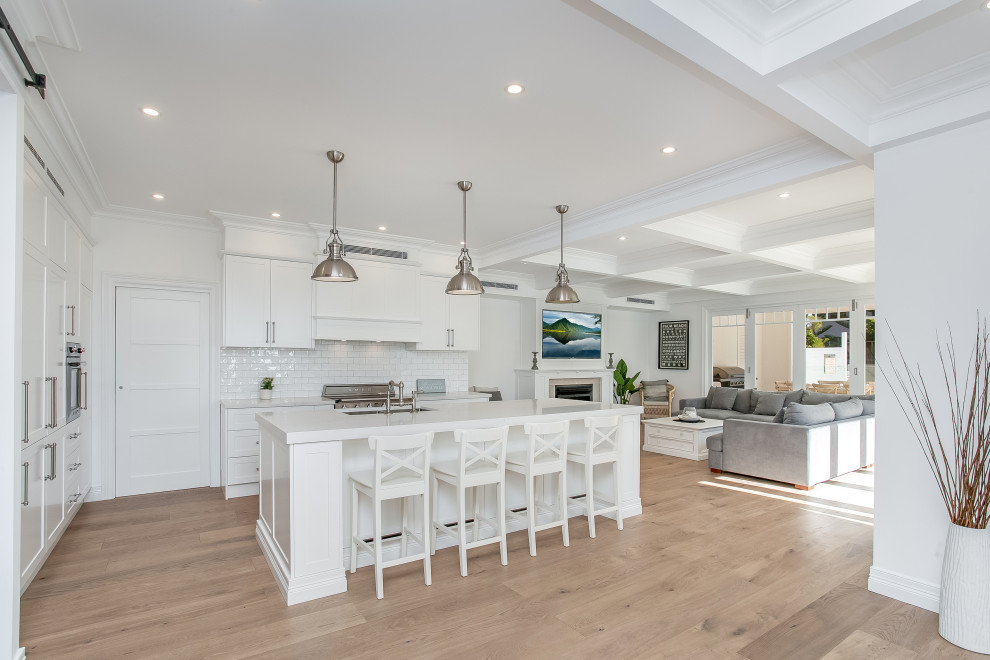 Photo of a large beach style kitchen pantry in Sydney with an undermount sink, shaker cabinets, white cabinets, quartz benchtops, white splashback, ceramic splashback, stainless steel appliances, medium hardwood floors, with island, brown floor, grey benchtop and coffered.