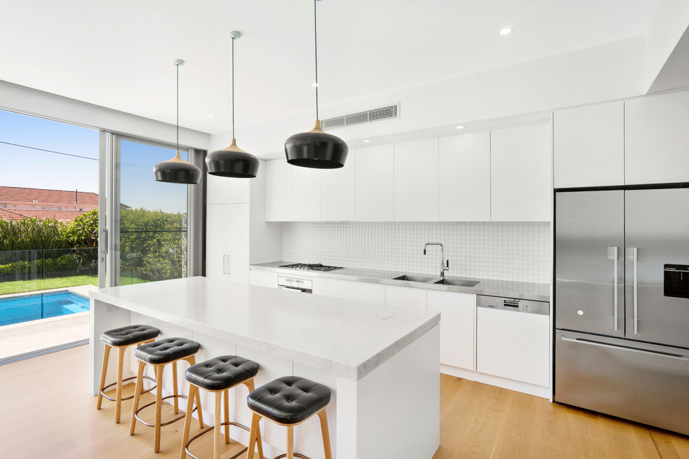 This is an example of a mid-sized contemporary single-wall open plan kitchen in Sydney with an integrated sink, flat-panel cabinets, stainless steel benchtops, white splashback, stainless steel appliances, medium hardwood floors, with island and brown floor.