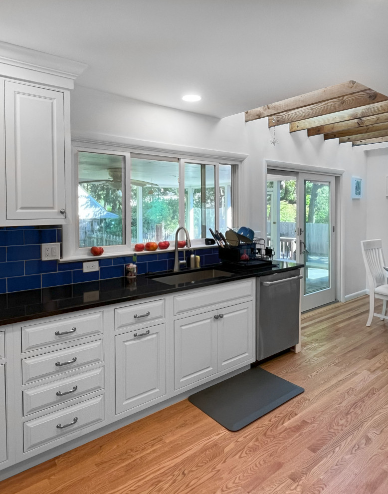 This is an example of a large beach style u-shaped kitchen/diner in Philadelphia with a submerged sink, raised-panel cabinets, engineered stone countertops, blue splashback, stainless steel appliances, medium hardwood flooring, brown floors and black worktops.