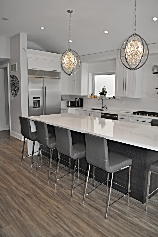 Photo of a mid-sized transitional l-shaped eat-in kitchen in Toronto with an undermount sink, shaker cabinets, white cabinets, quartz benchtops, white splashback, stone slab splashback, stainless steel appliances, vinyl floors, with island, brown floor and white benchtop.