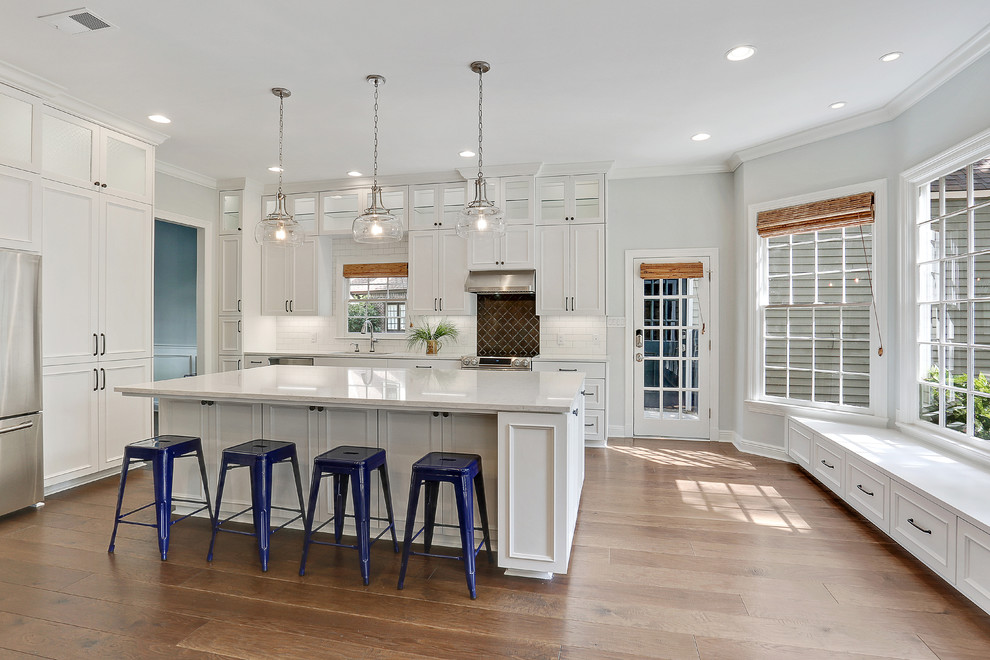 Photo of a mid-sized transitional l-shaped open plan kitchen in New Orleans with an undermount sink, white cabinets, quartz benchtops, white splashback, subway tile splashback, stainless steel appliances, with island, brown floor, white benchtop, recessed-panel cabinets and medium hardwood floors.