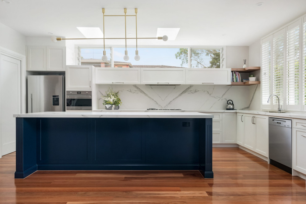 This is an example of a transitional l-shaped kitchen pantry in Sydney with a double-bowl sink, shaker cabinets, blue cabinets, white splashback, engineered quartz splashback, stainless steel appliances, medium hardwood floors, with island, white benchtop and tile benchtops.