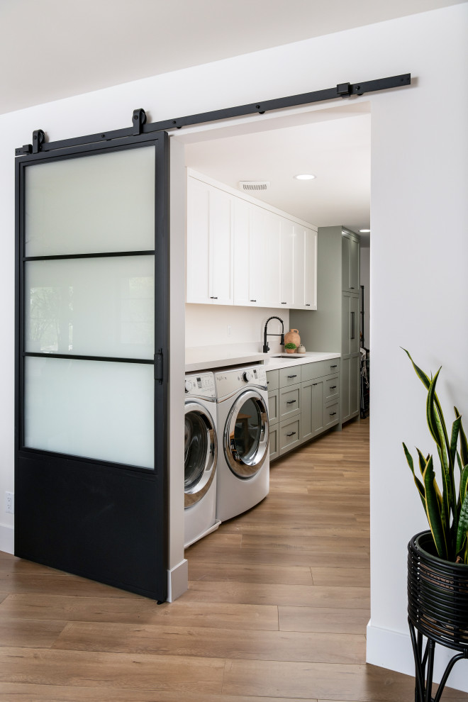 Photo of a mid-sized beach style galley dedicated laundry room in Orange County with a drop-in sink, shaker cabinets, green cabinets, quartz benchtops, white splashback, engineered quartz splashback, white walls, medium hardwood floors, a side-by-side washer and dryer and white benchtop.