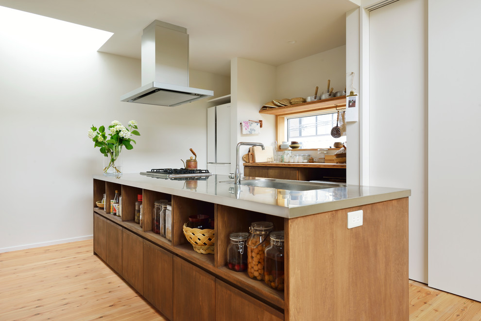 Photo of an asian galley kitchen in Tokyo with an integrated sink, flat-panel cabinets, medium wood cabinets, stainless steel benchtops, white splashback, light hardwood floors, with island, beige floor and grey benchtop.