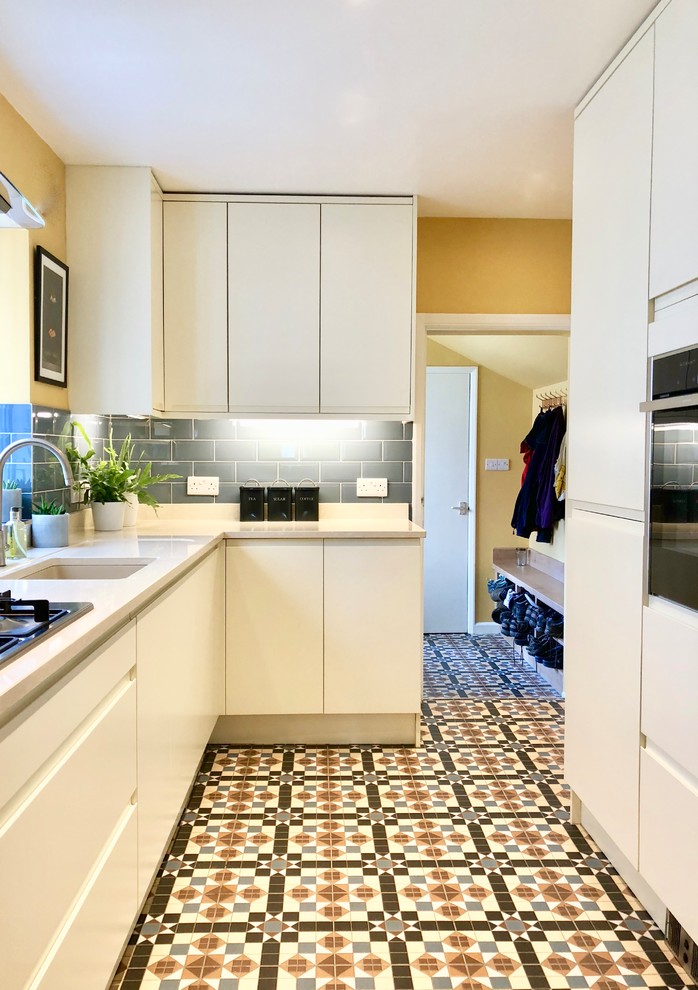 Photo of a mid-sized traditional galley separate kitchen in Other with an undermount sink, flat-panel cabinets, beige cabinets, solid surface benchtops, blue splashback, ceramic splashback, stainless steel appliances, ceramic floors, no island, multi-coloured floor and beige benchtop.