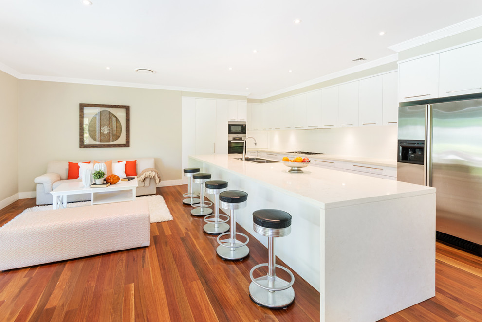 Large contemporary l-shaped open plan kitchen in Sydney with a double-bowl sink, flat-panel cabinets, white cabinets, white splashback, glass sheet splashback, stainless steel appliances, medium hardwood floors and with island.