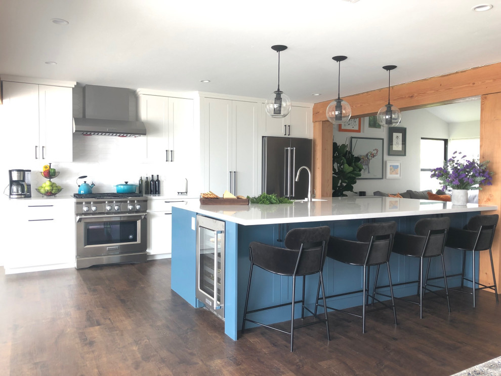 This is an example of an expansive country galley open plan kitchen in San Diego with a belfast sink, shaker cabinets, blue cabinets, quartz worktops, white splashback, porcelain splashback, stainless steel appliances, vinyl flooring, an island, brown floors, white worktops and exposed beams.