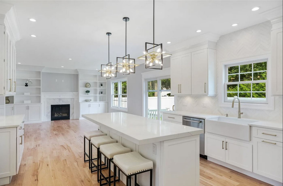 Large transitional u-shaped open plan kitchen in New York with a farmhouse sink, shaker cabinets, white cabinets, solid surface benchtops, white splashback, marble splashback, stainless steel appliances, medium hardwood floors, with island, brown floor and white benchtop.