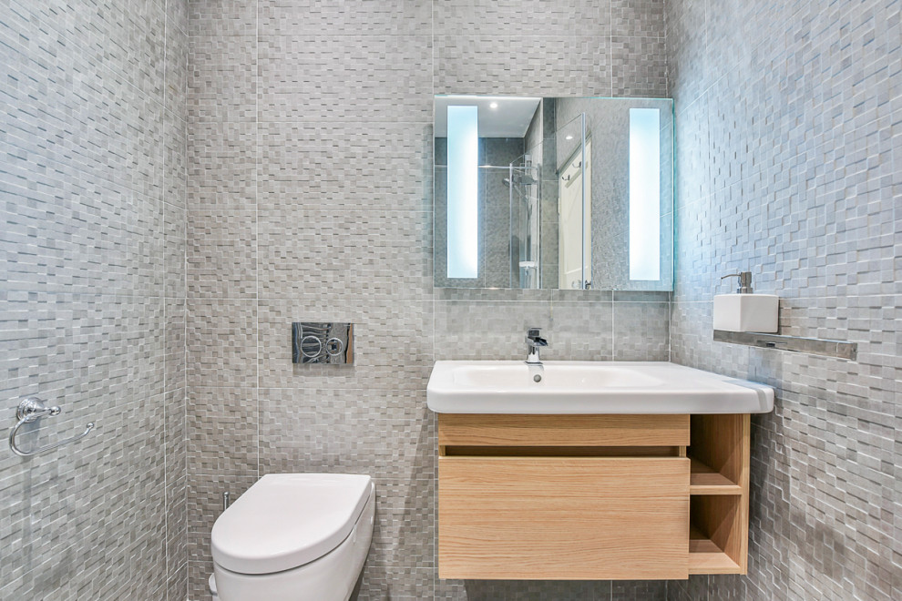 This is an example of a small contemporary kids wet room bathroom in London with flat-panel cabinets, light wood cabinets, a wall-mount toilet, gray tile, ceramic tile, grey walls, porcelain floors, a wall-mount sink, grey floor and a hinged shower door.