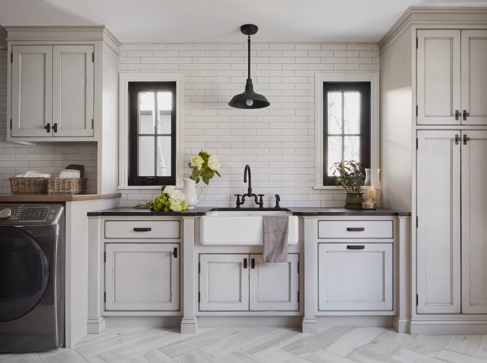 Inspiration for a large country single-wall dedicated laundry room in Denver with a farmhouse sink, recessed-panel cabinets, grey cabinets, soapstone benchtops, white walls, porcelain floors, a side-by-side washer and dryer, grey floor and black benchtop.