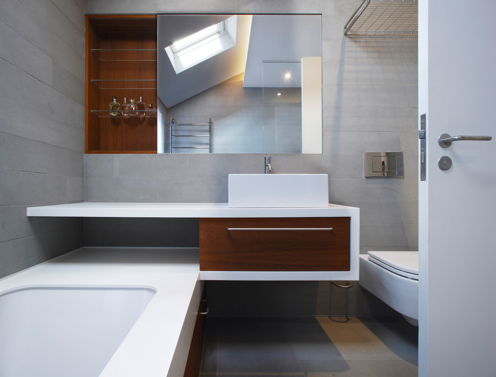 Inspiration for a mid-sized contemporary kids bathroom in London with open cabinets, brown cabinets, a drop-in tub, an open shower, a wall-mount toilet, gray tile, stone tile, grey walls, slate floors, a drop-in sink, solid surface benchtops, grey floor and an open shower.