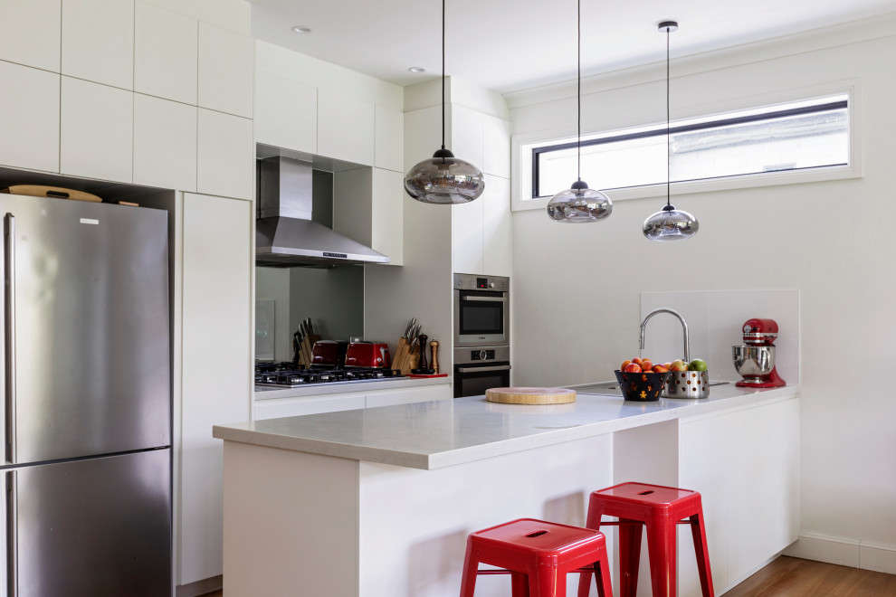 Contemporary galley kitchen in Sydney with flat-panel cabinets, white cabinets, stainless steel appliances, medium hardwood floors, a peninsula, brown floor and white benchtop.