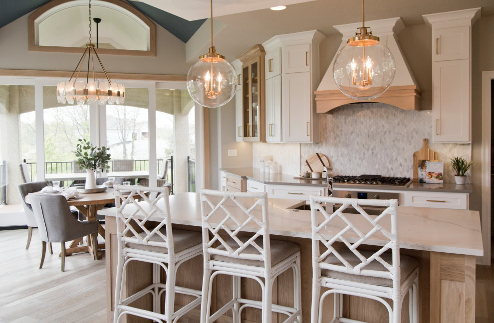 Photo of a large beach style l-shaped open plan kitchen in Kansas City with a farmhouse sink, shaker cabinets, light wood cabinets, quartz benchtops, white splashback, ceramic splashback, stainless steel appliances, light hardwood floors, with island, beige floor and white benchtop.