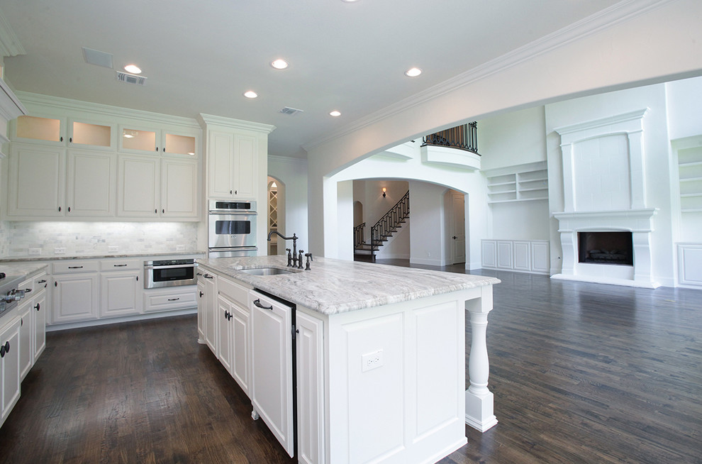 Large transitional l-shaped open plan kitchen in Dallas with a single-bowl sink, raised-panel cabinets, white cabinets, granite benchtops, grey splashback, stainless steel appliances, medium hardwood floors and with island.