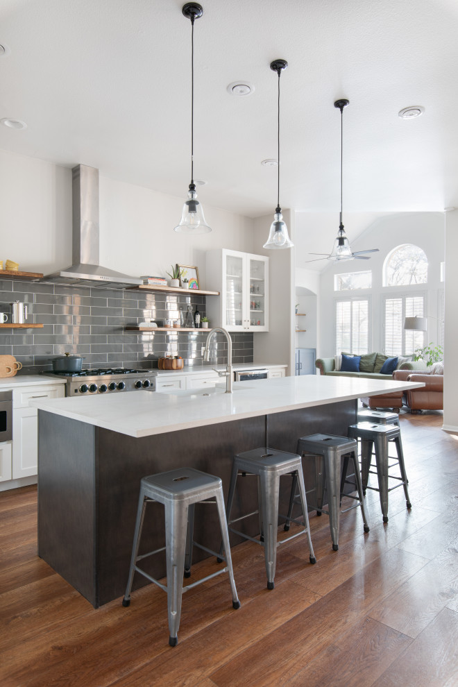 Photo of a large transitional l-shaped open plan kitchen in Austin with a farmhouse sink, shaker cabinets, white cabinets, quartz benchtops, grey splashback, subway tile splashback, stainless steel appliances, medium hardwood floors, with island, brown floor and white benchtop.
