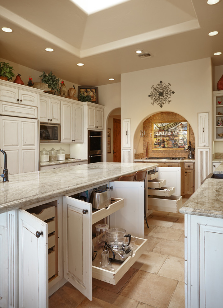 This is an example of a large country u-shaped eat-in kitchen in Other with a farmhouse sink, raised-panel cabinets, distressed cabinets, granite benchtops, beige splashback, panelled appliances, travertine floors, with island and beige floor.