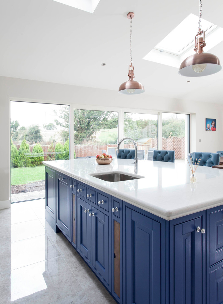 Expansive transitional l-shaped open plan kitchen in Dublin with an integrated sink, louvered cabinets, blue cabinets, quartzite benchtops, white splashback, stone slab splashback, stainless steel appliances, ceramic floors and with island.