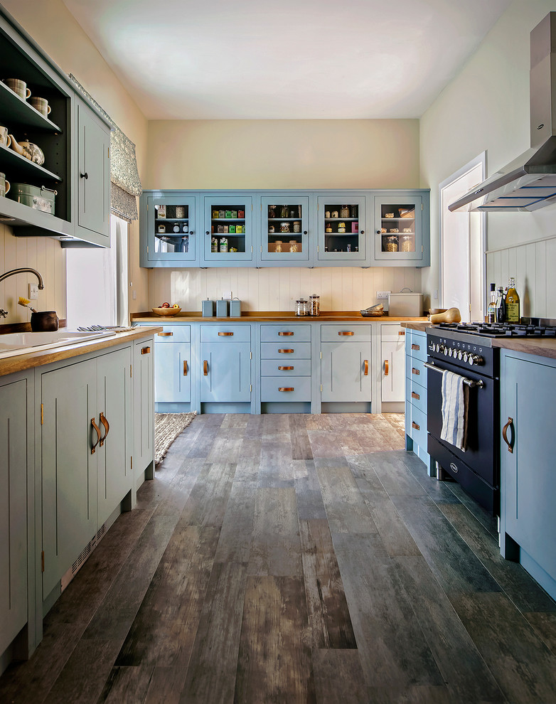 Traditional galley separate kitchen in London with a drop-in sink, shaker cabinets, blue cabinets, wood benchtops, beige splashback, black appliances, medium hardwood floors, no island and brown floor.