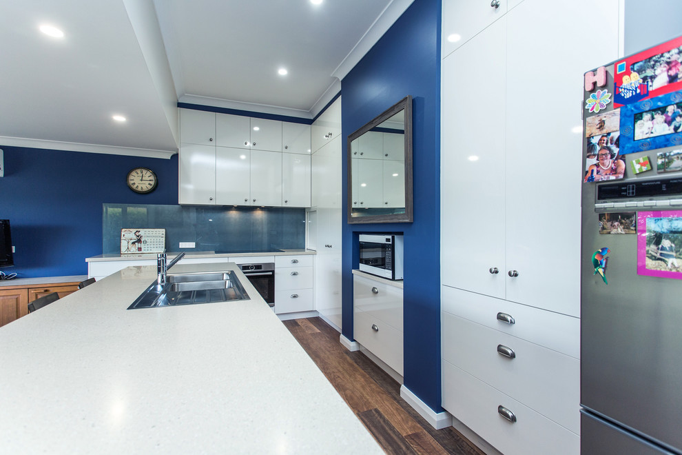 Photo of a small contemporary galley open plan kitchen in Other with a drop-in sink, flat-panel cabinets, white cabinets, solid surface benchtops, blue splashback, glass sheet splashback, stainless steel appliances, medium hardwood floors, a peninsula, brown floor and white benchtop.