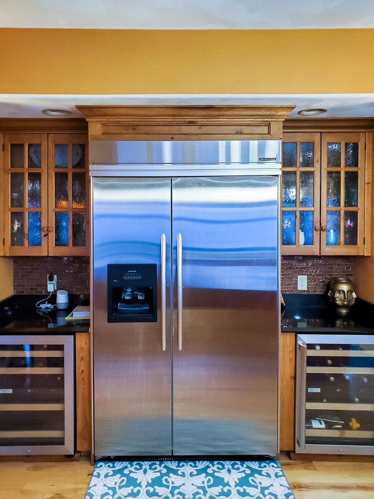 Eat-in kitchen - large rustic single-wall light wood floor and beige floor eat-in kitchen idea in Chicago with glass-front cabinets, medium tone wood cabinets, marble countertops, brown backsplash, stone tile backsplash, stainless steel appliances, an island and black countertops