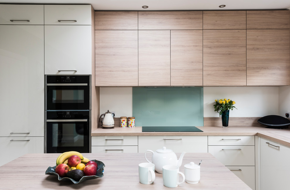 Photo of a scandinavian l-shaped eat-in kitchen in Surrey with a single-bowl sink, flat-panel cabinets, light wood cabinets, laminate benchtops, green splashback, glass sheet splashback, black appliances, ceramic floors, with island, beige floor and beige benchtop.