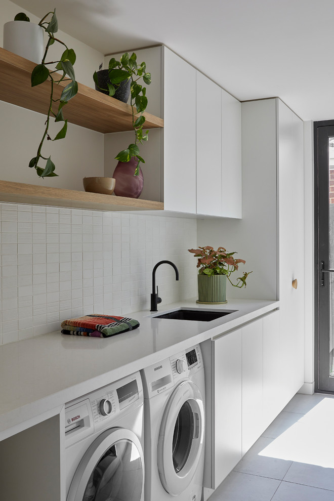 Zweizeilige, Mittelgroße Moderne Waschküche mit Unterbauwaschbecken, weißen Schränken, Quarzwerkstein-Arbeitsplatte, Küchenrückwand in Weiß, Rückwand aus Mosaikfliesen, weißer Wandfarbe, Waschmaschine und Trockner nebeneinander, grauem Boden und weißer Arbeitsplatte in Melbourne