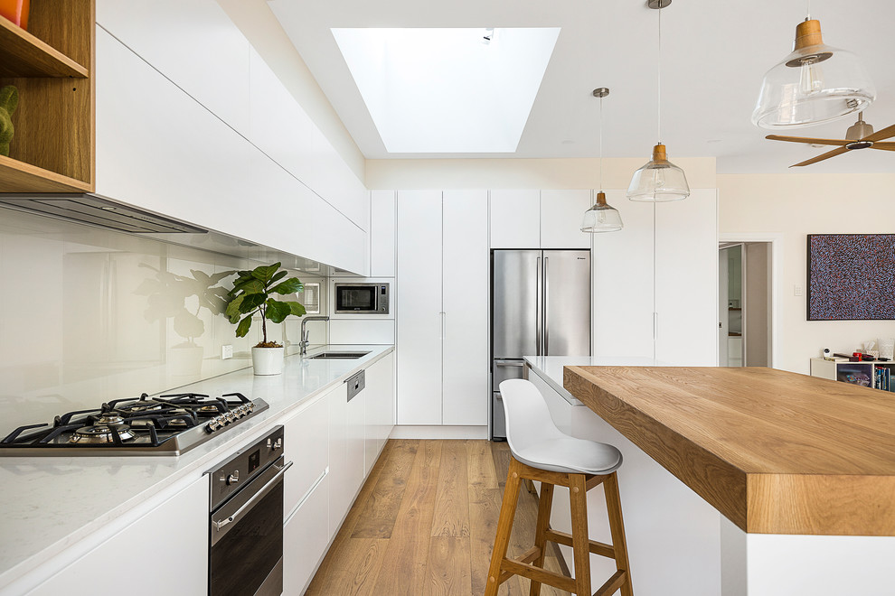 Inspiration for a large scandinavian l-shaped open plan kitchen in Sydney with an undermount sink, flat-panel cabinets, white cabinets, quartz benchtops, white splashback, glass sheet splashback, stainless steel appliances, medium hardwood floors, with island, white benchtop and brown floor.