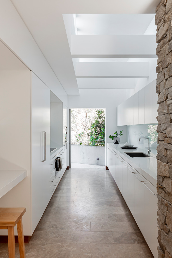 This is an example of a large midcentury galley separate kitchen in Sydney with an undermount sink, quartz benchtops, white splashback, glass sheet splashback, black appliances, travertine floors, brown floor and white benchtop.