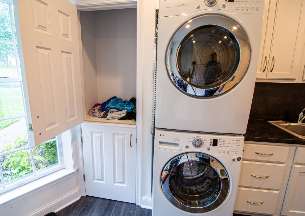 This is an example of a large traditional single-wall utility room in Philadelphia with a single-bowl sink, flat-panel cabinets, white cabinets, laminate benchtops, white walls, vinyl floors, a stacked washer and dryer, black floor and black benchtop.