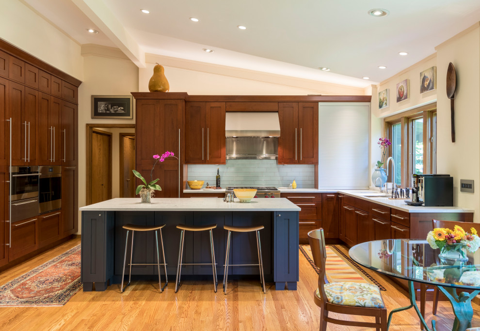 This is an example of a mid-sized transitional u-shaped open plan kitchen in St Louis with an undermount sink, shaker cabinets, brown cabinets, quartz benchtops, grey splashback, glass tile splashback, panelled appliances, light hardwood floors, with island, brown floor, white benchtop and vaulted.
