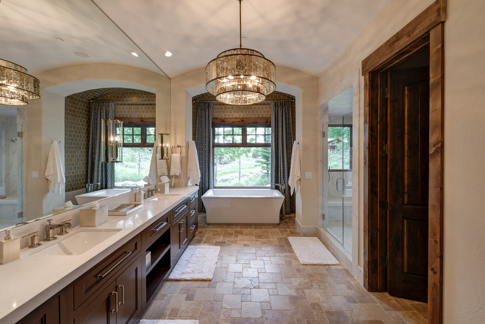 This is an example of a large mediterranean master bathroom in Salt Lake City with recessed-panel cabinets, a freestanding tub, beige walls, travertine floors, engineered quartz benchtops, a hinged shower door, white benchtops, dark wood cabinets, an alcove shower, an undermount sink and brown floor.