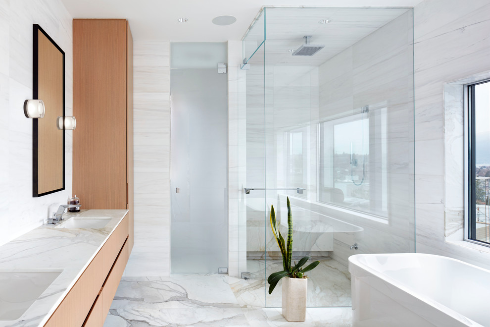 This is an example of a large contemporary master bathroom in Vancouver with flat-panel cabinets, light wood cabinets, a freestanding tub, a corner shower, white tile, stone tile, white walls, marble floors, an undermount sink, marble benchtops and a hinged shower door.