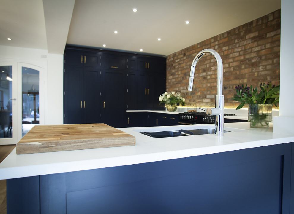 Design ideas for a mid-sized industrial l-shaped open plan kitchen in Other with an undermount sink, shaker cabinets, black cabinets, solid surface benchtops, brick splashback, black appliances, light hardwood floors, no island, beige floor and white benchtop.