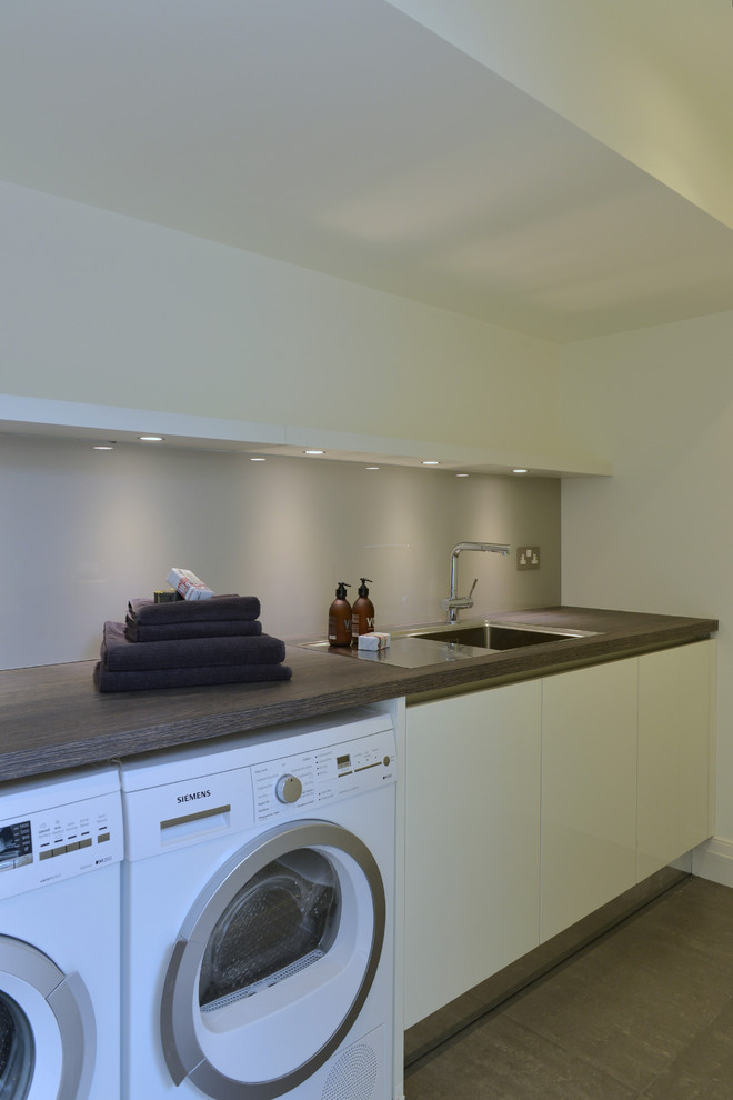Photo of a small contemporary l-shaped dedicated laundry room in Gloucestershire with flat-panel cabinets, white cabinets, laminate benchtops, white walls, porcelain floors, a stacked washer and dryer and a drop-in sink.