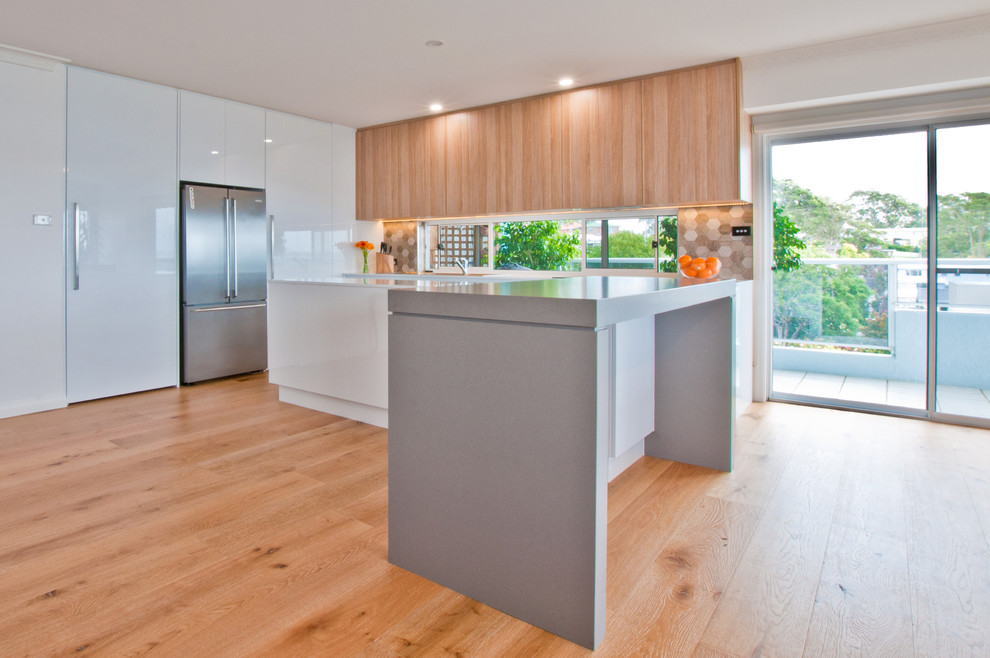 Photo of a mid-sized modern l-shaped open plan kitchen in Sydney with a drop-in sink, flat-panel cabinets, light wood cabinets, quartzite benchtops, grey splashback, stone tile splashback, stainless steel appliances, light hardwood floors and with island.