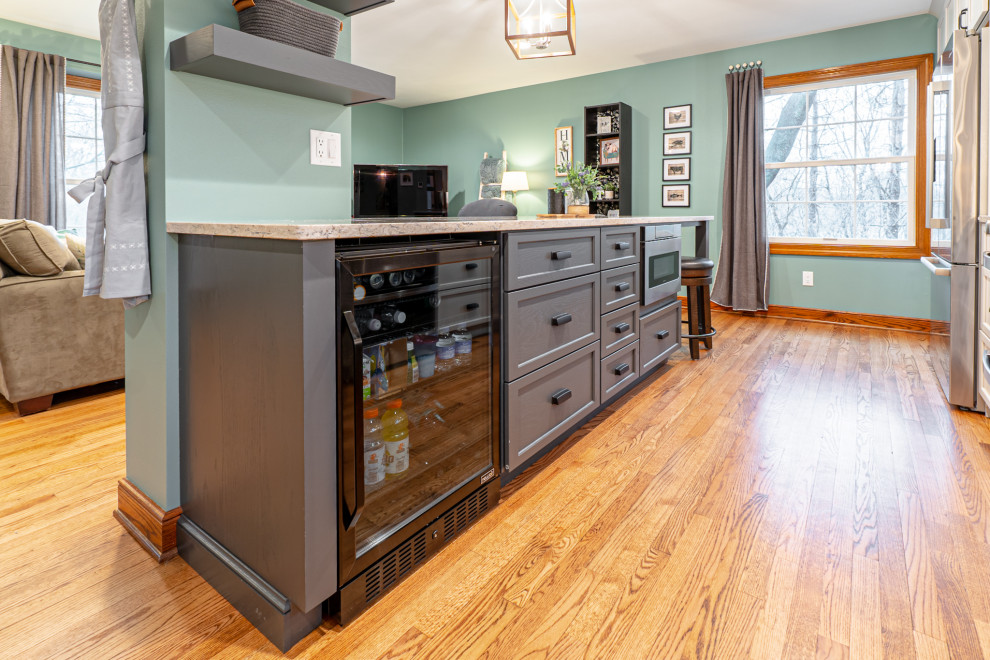 This is an example of a mid-sized country galley open plan kitchen in Milwaukee with a farmhouse sink, shaker cabinets, beige cabinets, quartz benchtops, white splashback, mosaic tile splashback, stainless steel appliances, medium hardwood floors, a peninsula, brown floor and multi-coloured benchtop.