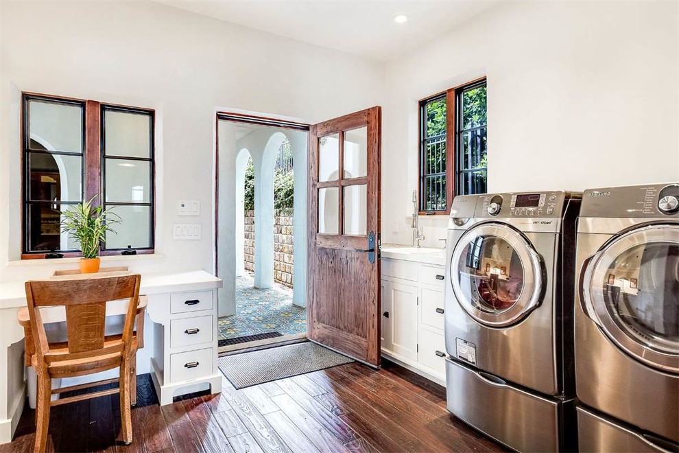 Mid-sized mediterranean l-shaped utility room in Austin with an integrated sink, white cabinets, white walls, dark hardwood floors, a side-by-side washer and dryer and brown floor.