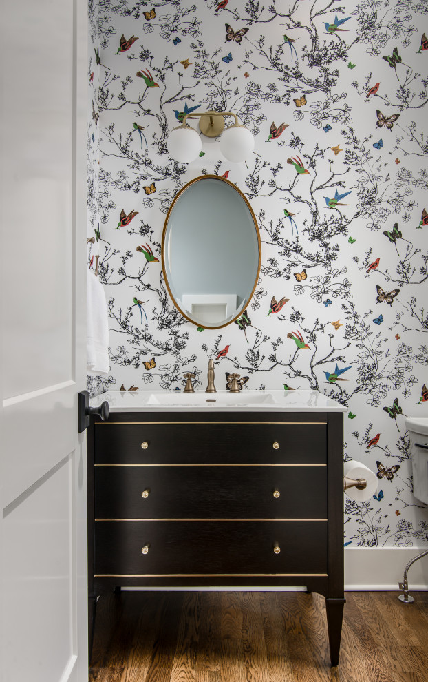 Small transitional powder room in Nashville with furniture-like cabinets, dark wood cabinets, a two-piece toilet, an integrated sink, marble benchtops, white benchtops, a freestanding vanity, wallpaper, multi-coloured walls, medium hardwood floors and brown floor.