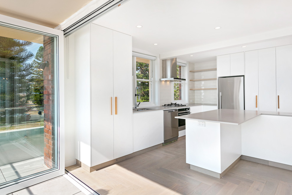 Design ideas for a mid-sized beach style galley open plan kitchen in Sydney with a double-bowl sink, flat-panel cabinets, white cabinets, quartz benchtops, white splashback, subway tile splashback, stainless steel appliances, light hardwood floors, with island, grey floor and grey benchtop.